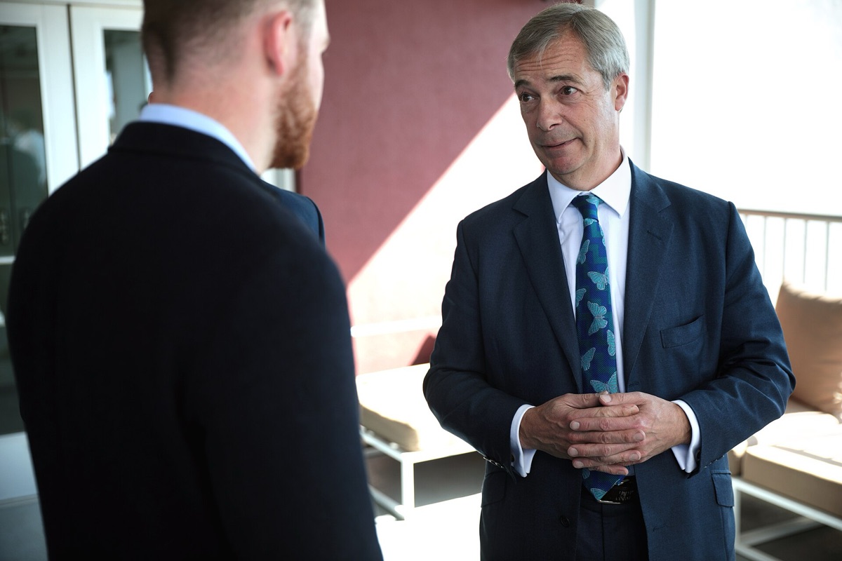 Nigel Farage speaking at event, crowd in background, neutral expression, outdoor setting